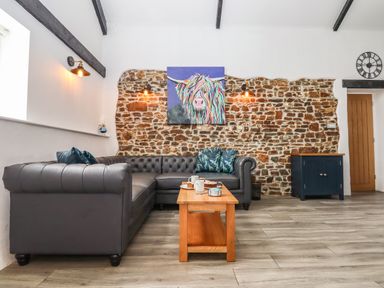 A living room with a gray corner sofa a wooden coffee table with mugs and plates exposed stone wall with colorful cow painting and wall lights at Strawberry Bank - Kingfisher in Pyworthy near Bridgerule