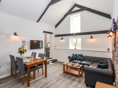 A living room with a dining table, chairs, a sofa, coffee table, TV on the wall, and exposed beams at Strawberry Bank - Kingfisher Pyworthy near Bridgerule