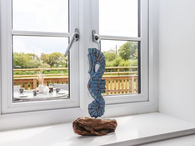 A blue seahorse sculpture on a wooden base on a white window sill with a view of a table and chairs outside at Strawberry Bank - Kingfisher Pyworthy near Bridgerule