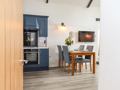 A kitchen and dining area with blue cabinets a wooden table with chairs and a wall-mounted television at Strawberry Bank - Kingfisher in Pyworthy near Bridgerule