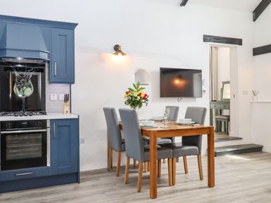 A kitchen with blue cabinets a wooden dining table with grey chairs and a wall-mounted TV at Strawberry Bank - Kingfisher in Pyworthy near Bridgerule