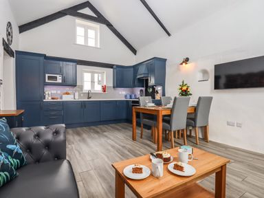 A kitchen and dining area with blue cabinets a wooden dining table with chairs and a coffee table with cake and mugs at Strawberry Bank - Kingfisher Pyworthy near Bridgerule