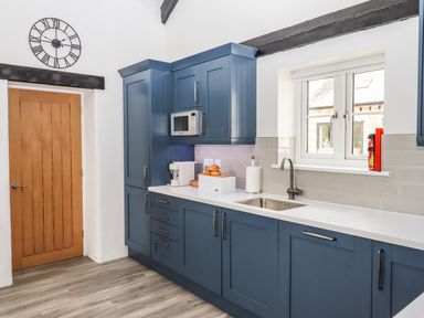 A kitchen with blue cabinets a wooden door a clock on the wall and a window over the sink at Strawberry Bank - Kingfisher Pyworthy near Bridgerule