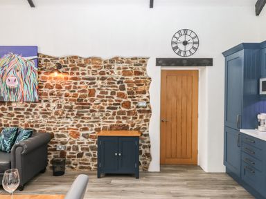 An interior room with a section of exposed brick wall a wooden door a black clock blue cabinets and a black leather sofa at Strawberry Bank - Kingfisher in Pyworthy near Bridgerule