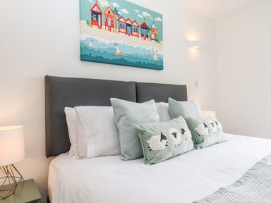 A bedroom with a bed with gray headboard and pillows with sheep designs and a seaside painting on the wall at Strawberry Bank Kingfisher in Pyworthy near Bridgerule