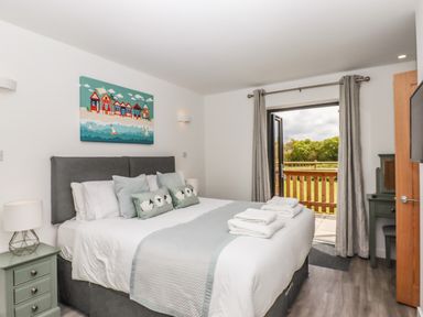 A bedroom with a large bed, bedside table with lamp, wall art above the bed, open door leading to a balcony with a view of greenery at Strawberry Bank - Kingfisher in Pyworthy near Bridgerule