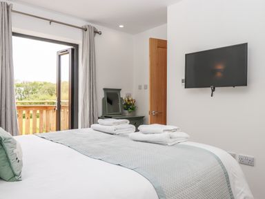 A bedroom with a bed, folded towels, a wall-mounted TV, open door, curtains, and a balcony at Strawberry Bank - Kingfisher in Pyworthy near Bridgerule