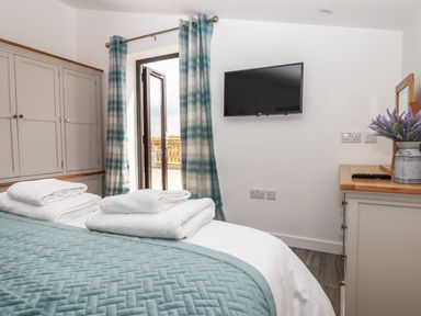 A bedroom with a bed covered by a blue quilt and stacked towels a wall-mounted tv curtains and a cabinet at Strawberry Bank - Kingfisher in Pyworthy near Bridgerule