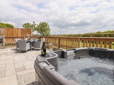 An outdoor patio with a hot tub and a dining table set with chairs and a bottle of prosecco at Strawberry Bank - Kingfisher in Pyworthy near Bridgerule