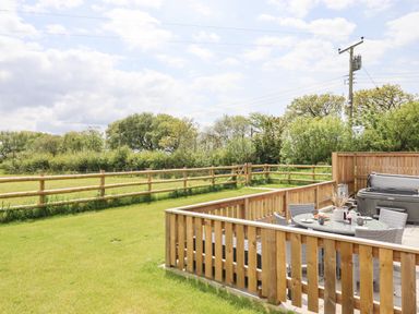 An outdoor fenced patio area with a table and chairs surrounded by a grassy field at Strawberry Bank - Kingfisher in Pyworthy near Bridgerule