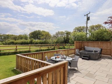 An outdoor patio with a round table and chairs next to a hot tub with wooden fencing around at Strawberry Bank - Kingfisher in Pyworthy near Bridgerule
