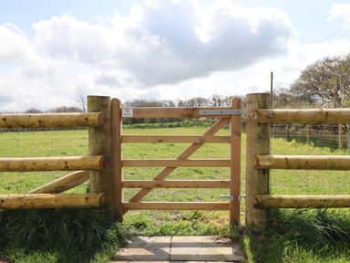 A wooden gate in a wooden fence opening to a green grassy field under a cloudy sky at Strawberry Bank - Kingfisher in Pyworthy near Bridgerule
