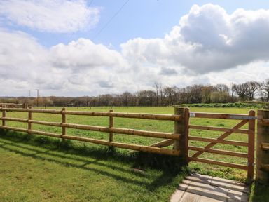 A wooden fence and gate enclosing a grassy field with trees in the background at Strawberry Bank - Kingfisher in Pyworthy near Bridgerule
