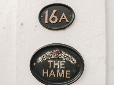 Black oval house number plaque with 16A above a black oval plaque with The Hame and floral decoration on a white wall at The Hame in Monmouth