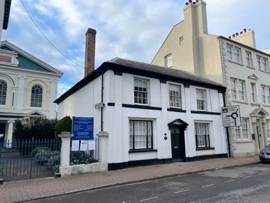 A street view showing a white building with black window frames and a blue sign outside next to a gated garden at The Hame in Monmouth