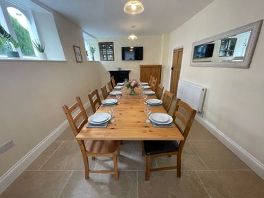 A dining room with a long wooden table set with plates and glasses surrounded by wooden chairs with plants on windowsills at The Hame in Monmouth