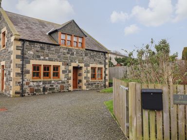A stone house with wooden framed windows a gravel driveway and a wooden gate at Whin Hill Cottage in Craster