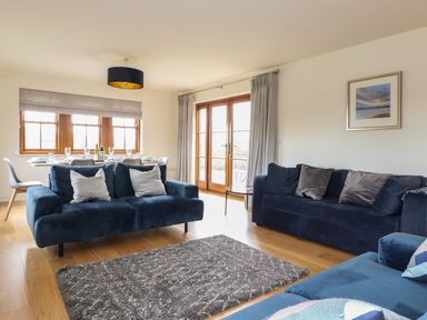 A living room with blue sofas a grey rug wooden floor and a dining table by windows at Whin Hill Cottage in Craster