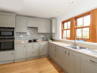 A kitchen with grey cabinets wooden floor double oven and a window with wooden frame at Whin Hill Cottage in Craster