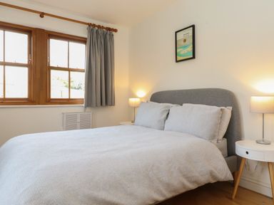 A bedroom with a bed gray headboard bedside tables lamps and windows with curtains at Whin Hill Cottage Craster