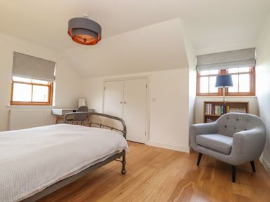 A bedroom with a bed desk chair armchair bookshelf and two windows at Whin Hill Cottage in Craster
