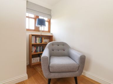 A grey armchair in front of a wooden bookshelf with books and a blue table lamp on top at Whin Hill Cottage in Craster