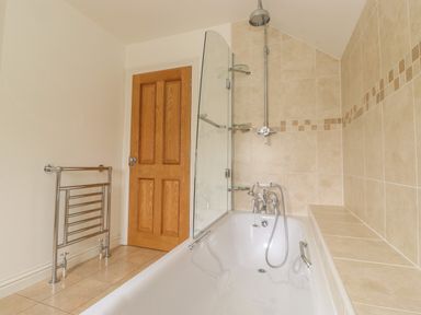 A bathroom with a bathtub shower combination tiled walls glass screen wooden door and a metal towel rail at Whin Hill Cottage in Craster