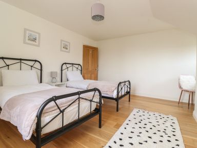 A bedroom with two single beds a nightstand with a lamp a door a chair with a cushion and a rug on a wooden floor at Whin Hill Cottage in Craster