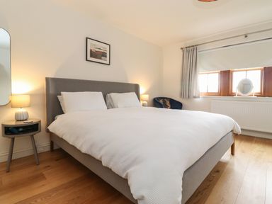 A bedroom with a double bed white bedding bed side tables lamps window with curtains and a blue chair at Whin Hill Cottage in Craster