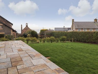 A garden with a stone patio lawn and hedges with houses in the background at Whin Hill Cottage in Craster