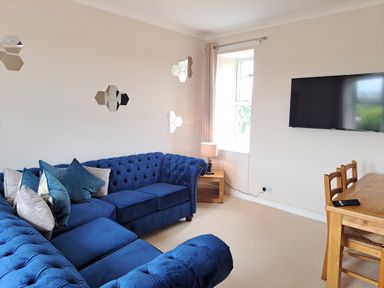 A living room with blue sectional sofa cushions TV on wall and a wooden dining table with chairs at Bogrie Cottage Canonbie near Gretna Green