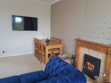 A dining table with chairs next to a wall mounted TV and a fireplace with a blue sofa in the foreground at Bogrie Cottage Canonbie near Gretna Green