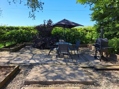 An outdoor patio area with a table and four chairs under an umbrella and a barbecue grill surrounded by greenery at Bogrie Cottage in Canonbie near Gretna Green