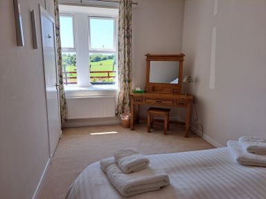 A bedroom with a bed, towels on the bed, a wooden dressing table with a mirror, a stool, a lamp, a window with curtains, and a door at Bogrie Cottage in Canonbie near Gretna Green