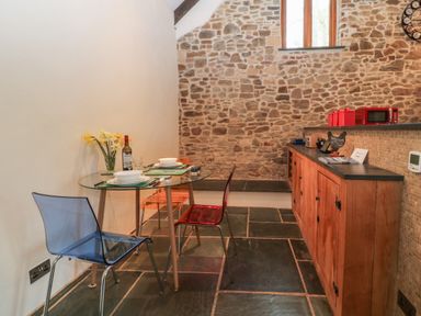 A dining area with a glass table set for two and colored chairs next to a wooden cabinet and stone wall at The Threshing Barn in Highampton
