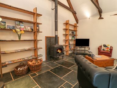 A living room with wooden shelves, a wood burning stove, a black leather sofa, a red armchair, a television, and a wooden coffee table at The Threshing Barn in Highampton
