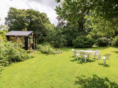 A garden with a black wooden shed and white plastic chairs around a table on the grass at The Threshing Barn in Highampton