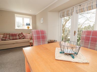 A dining table with a wire basket and chairs beside a sofa with plaid cushions in a living room with windows and a door at Magnolia House in Dittisham