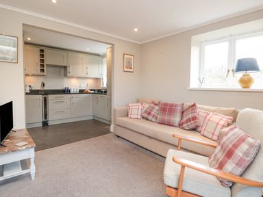 A living room with a beige sofa and plaid cushions next to a window with a lamp and an armchair adjacent to a kitchen with cabinets and appliances at Magnolia House in Dittisham