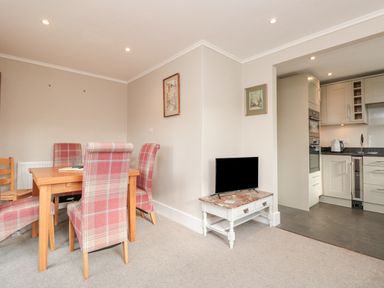 A dining area with a wooden table and plaid chairs next to a small white table with a TV and an open kitchen at Magnolia House in Dittisham