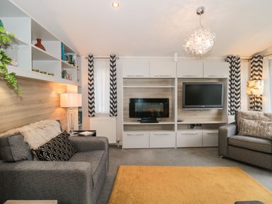 A living room with two gray sofas a wall unit with a television and electric fireplace and patterned curtains at Owls Nest in Furzehill