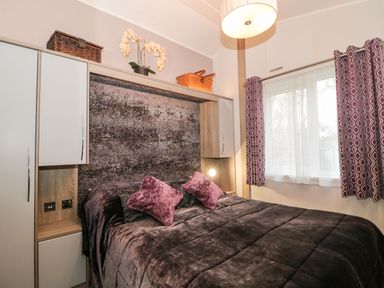 A bedroom with a bed with purple cushions and a textured headboard under cabinets and a window with curtains at Owls Nest in Furzehill