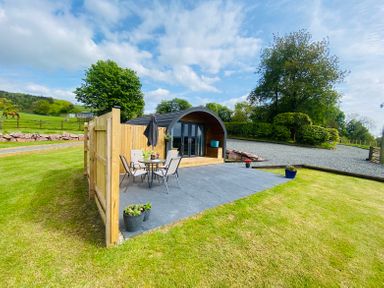 A small wooden pod with glass doors and patio furniture on a slate patio in a grassy area at Dryfe Lodge in Lockerbie