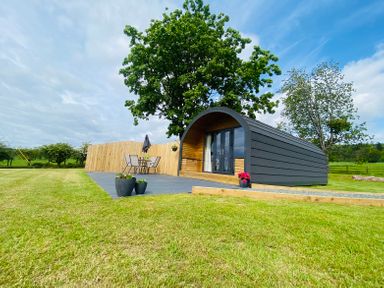 A small curved cabin with glass doors on a tiled patio with chairs and table surrounded by grass and trees at Dryfe Lodge in Lockerbie