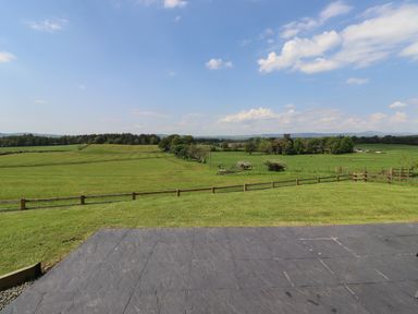 A garden with a paved patio and fenced green fields under a blue sky at Dryfe Lodge in Lockerbie