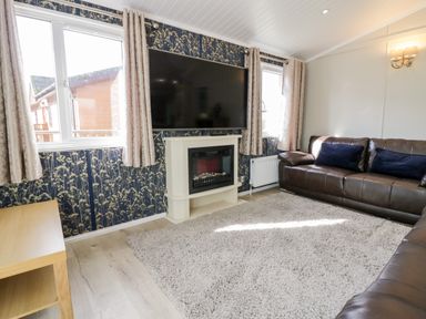 A living room with a large wall-mounted TV a white fireplace two windows brown leather sofas and a light rug at Lake View Lodge in Lakeside 3 White Cross Bay Windermere