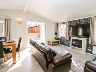 A living room with brown leather sofas a large wall-mounted TV a fireplace a wooden dining table with black chairs and an open door leading outside at Lake View Lodge in Lakeside 3 White Cross Bay Windermere