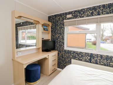 A bedroom corner with a wooden dressing table mirror drawers a blue stool beside a window with floral wallpaper at Lake View Lodge Lakeside 3 White Cross Bay Windermere