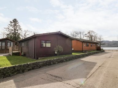 Two wooden lodges beside a stone wall and a paved road with a lake and trees in the background at Lake View Lodge in Lakeside 3 White Cross Bay Windermere