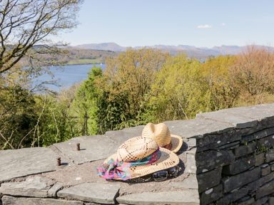 A stone wall with two straw hats and sunglasses overlooking trees and a lake with hills in the distance at Lake View Lodge Lakeside 3 White Cross Bay Windermere
